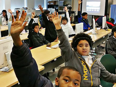 Children seated in a classroom with their hands raised. 