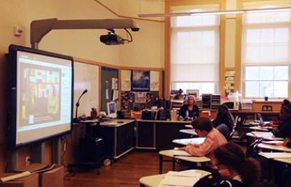 Children seated at desks in a classroom in front of a projector. 